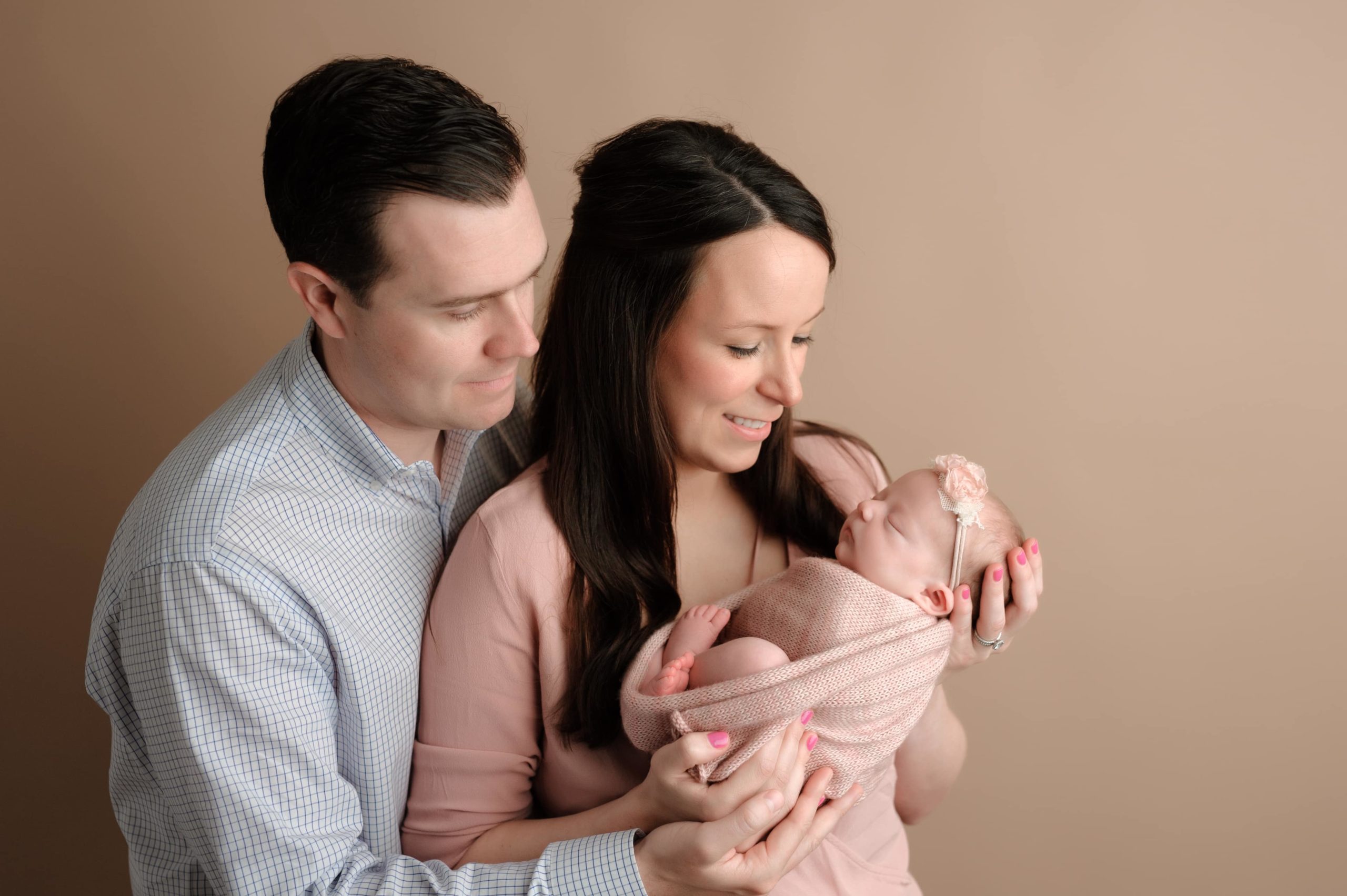 Newborn photoshoot with mom and dad holding their baby in front of a deep beige background