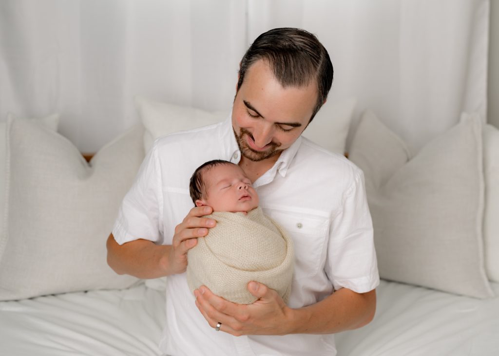 Dad in white shirt holds swaddled newborn and smiles down at the baby