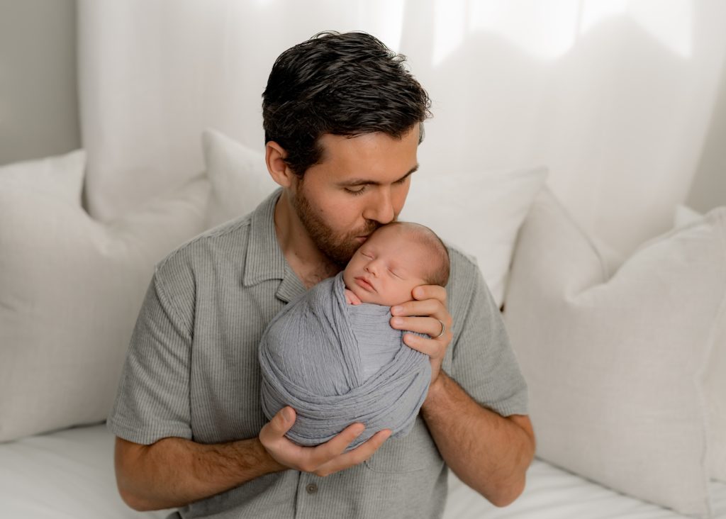 Dad holds swaddled newborn, kissing him on cheek